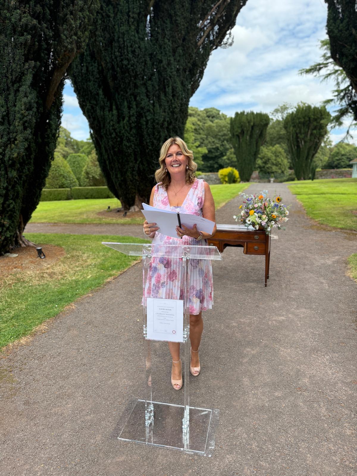 Grá delivering a ceremony at a glass lectern in a garden lined with cypress trees